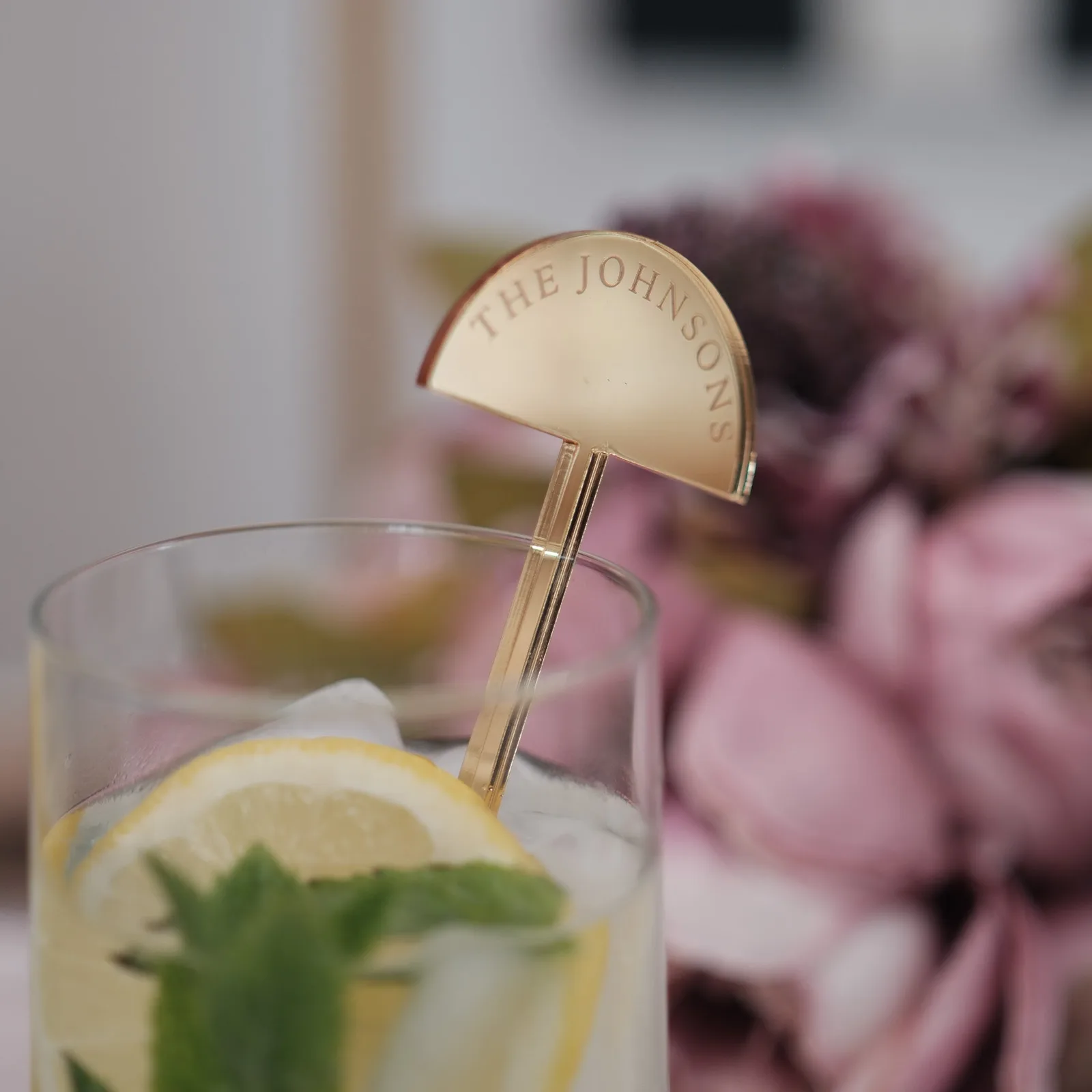 Mirror gold half-moon stirrer engraved with a family name, standing in a highball with lemon and mint